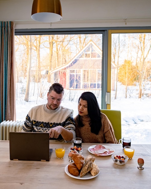Inside a brightly lit tiny house, a young couple is preparing a meal in a compact yet fully functional kitchen. The space is efficiently organized with custom cabinetry, stainless steel appliances, and a small dining area. The large windows offer a view of the American countryside, creating a warm and inviting atmosphere.