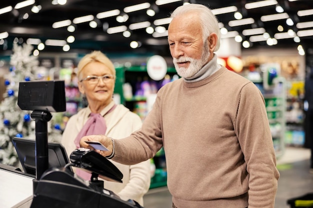 A senior man showing his senior discount card at a store checkout, with the cashier smiling and pointing to a discounted price tag on a popular gadget.
