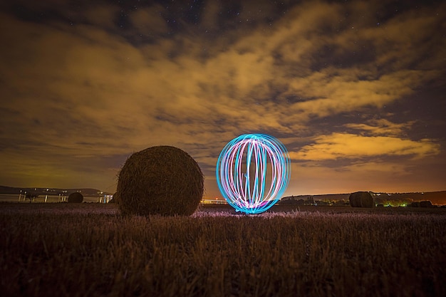 A night shot showing a crop circle in a field in Kansas. A beam of light shines down from the sky onto the crop circle, illuminating the intricate pattern. The stars are visible in the dark sky, and the overall scene has an ethereal quality.